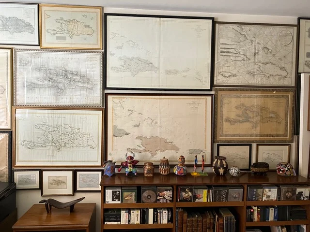  Vista parcial de la colección de mapas antiguos de Joseph González y Ralph Magnus, donde se destacan cartas náuticas y representaciones tempranas del Caribe y América Central. La disposición mural se complementa con una selección de porcelanas, lámpara y objetos decorativos sobre un aparador, creando un entorno que celebra la historia y el coleccionismo .