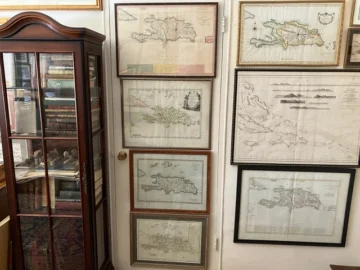  Detalle de la colección de Joseph González y Ralph Magnus, donde se aprecian mapas antiguos de La Española y otras islas del Caribe, acompañados por una vitrina con libros raros y objetos históricos.